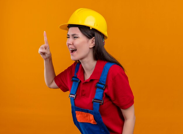 Annoyed young builder girl points up and looks to the side on isolated orange background
