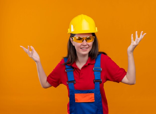 Annoyed young builder girl with safety glasses raised both hands up on isolated orange background