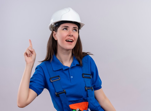 Annoyed young builder girl with white safety helmet and blue uniform points up and looks up on isolated white background with copy space