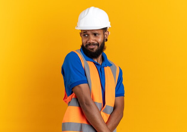 annoyed young builder man in uniform with safety helmet looking at side isolated on orange wall with copy space