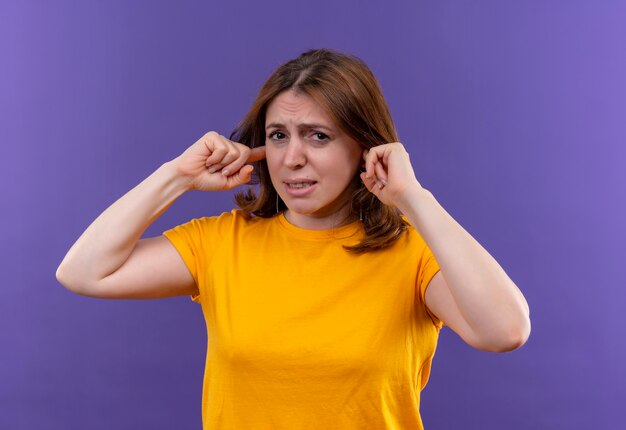 Раздраженная молодая случайная женщина кладет пальцы на уши на изолированном фиолетовом пространстве