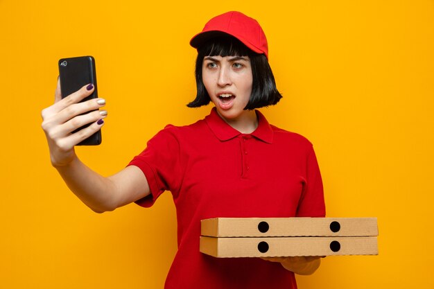 Annoyed young caucasian delivery girl holding pizza boxes and yelling at someone looking at phone 