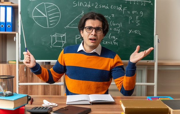 빈 손을 보여주는 앞을 바라보는 포인터 스틱을 들고 교실에서 학용품을 들고 책상에 앉아 안경을 쓴 젊은 백인 기하학 교사