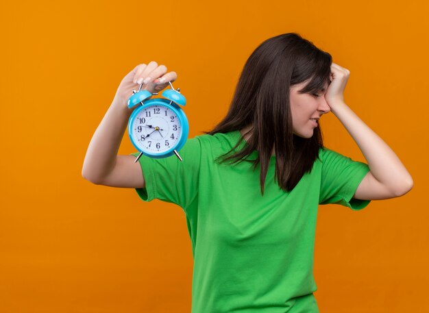 Annoyed young caucasian girl in green shirt holds clock and puts hand on head on isolated orange background