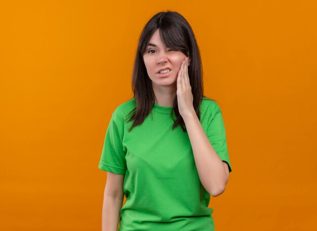 Annoyed young caucasian girl in green shirt holds face and looks at camera on isolated orange background