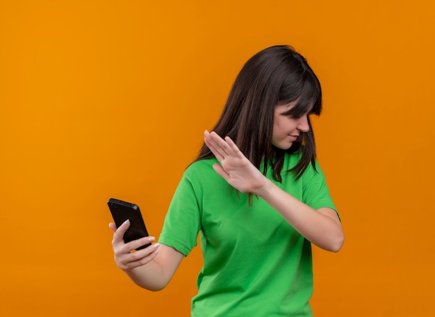 Foto gratuita la giovane ragazza caucasica infastidita in camicia verde tiene il telefono e gesti no su fondo arancio isolato con lo spazio della copia