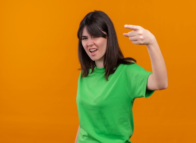 Annoyed young caucasian girl in green shirt points forward and looks at camera on isolated orange background with copy space