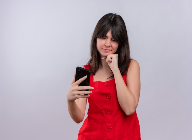 Annoyed young caucasian girl holding phone and keeping hand on chin looking at camera on isolated white background