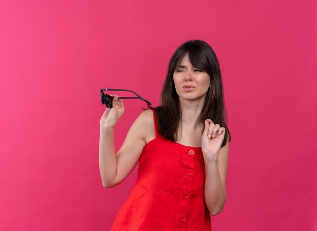 Annoyed young caucasian girl holding sunglasses and looking to the side on isolated pink background