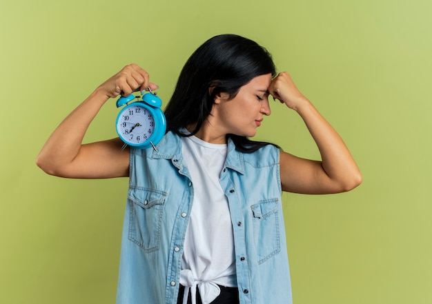 Annoyed young caucasian girl puts fist on forehead and holds alarm clock isolated on olive green background with copy space