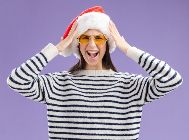 Annoyed young caucasian girl in sun glasses with santa hat holds head yelling at someone isolated on purple wall with copy space