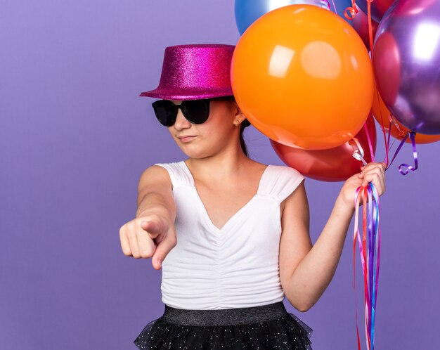 annoyed young caucasian girl in sun glasses with violet party hat holding helium balloons and pointing  isolated on purple wall with copy space