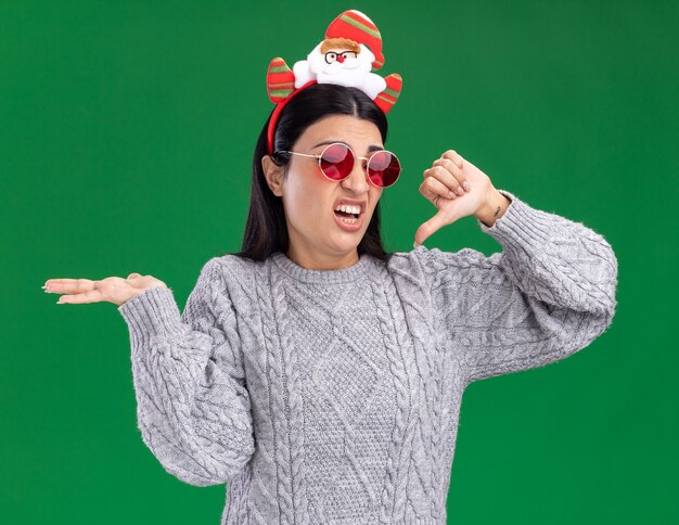 Annoyed young caucasian girl wearing santa claus headband with glasses looking at camera showing empty hand and thumb down isolated on green background