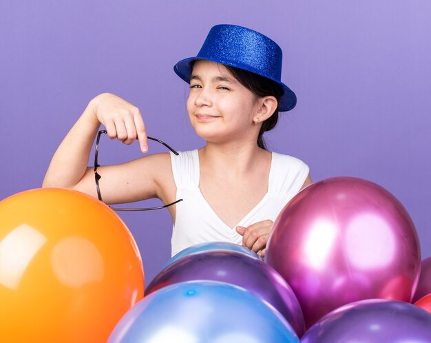 раздраженная молодая кавказская девушка в синей партийной шляпе, держащая оптические очки, стоя с гелиевыми шарами, изолированными на фиолетовой стене с копией пространства