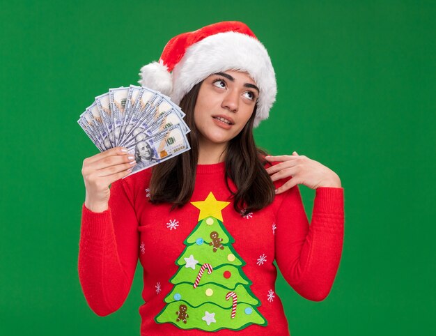 サンタの帽子をかぶったイライラする若い白人の女の子は、コピースペースで緑の背景に分離された側を見てお金を保持します