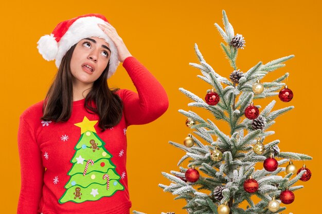 Annoyed young caucasian girl with santa hat puts hand on forehead rolling eyes standing next to christmas tree isolated on orange background with copy space