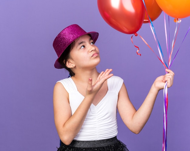 annoyed young caucasian girl with violet party hat holding and pointing at helium balloons isolated on purple wall with copy space