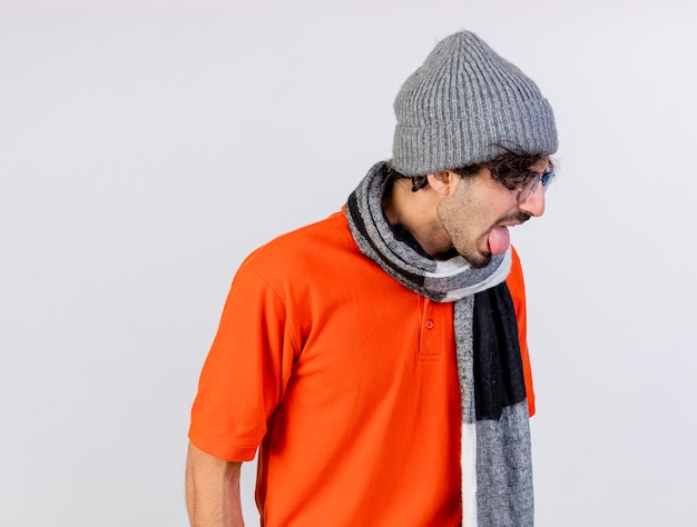 Infastidito giovane indoeuropeo uomo malato con gli occhiali inverno cappello e sciarpa girando testa a lato che mostra la lingua isolata sulla parete bianca con lo spazio della copia