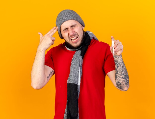 Annoyed young caucasian ill man wearing winter hat and scarf gestures gun hand sign