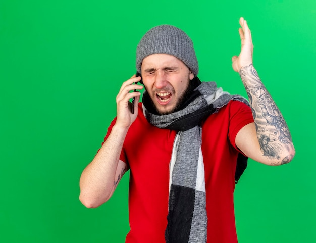 冬の帽子とスカーフを身に着けているイライラする若い白人の病気の男は、電話で話している手を上げて立っています