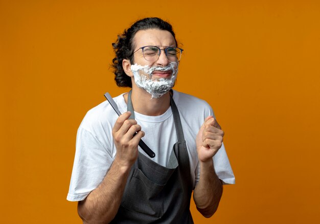 Annoyed young caucasian male barber wearing glasses and wavy hair band in uniform holding straight razor with shaving cream put on his face with closed eyes