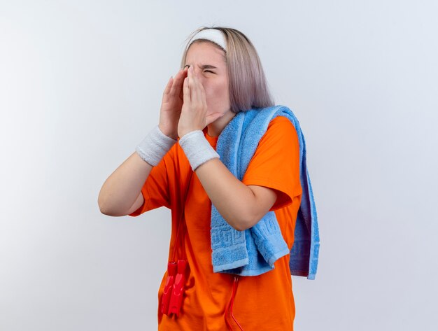 Раздраженная молодая кавказская спортивная девушка с подтяжками и со скакалкой на шее в браслетах с повязкой на голову держит полотенце на плече и зовет кого-то, смотрящего в сторону на белой стене