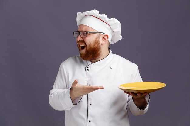 Annoyed young chef wearing glasses uniform and cap holding and pointing with hand at empty plate looking at side isolated on purple background