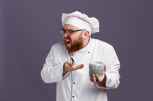 Annoyed young chef wearing glasses uniform and cap holding spoon and bowl looking at side pointing at bowl isolated on purple background