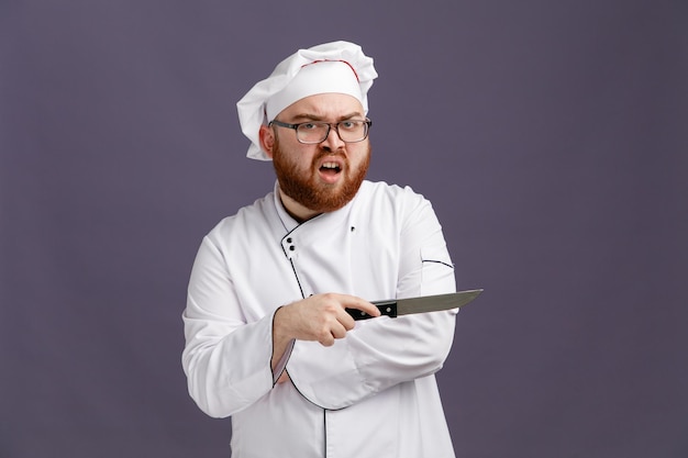 Annoyed young chef wearing glasses uniform and cap looking at camera pointing to side with knife isolated on purple background