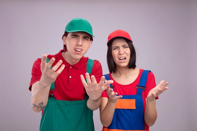 Annoyed young couple in construction worker uniform and cap looking at camera showing empty hands isolated on white wall