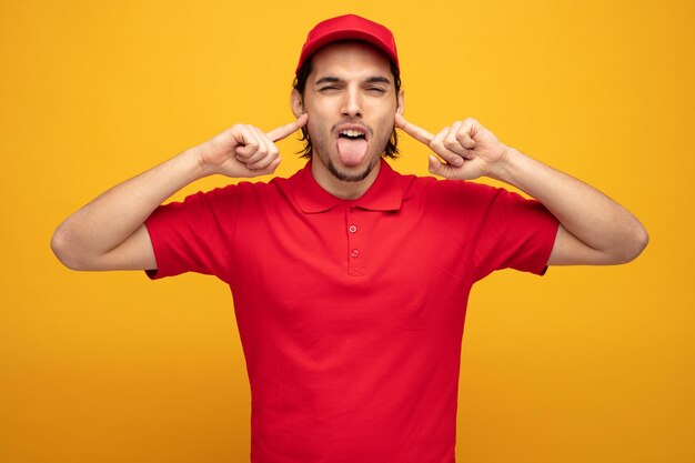 annoyed young delivery man wearing uniform and cap putting fingers in ears looking at camera showing tongue isolated on yellow background