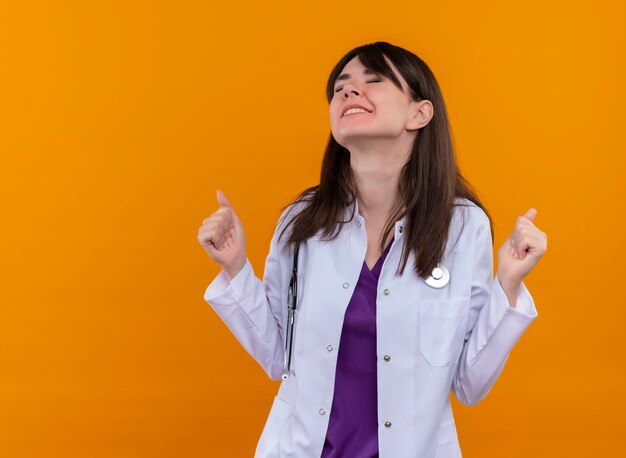 Annoyed young female doctor in medical robe with stethoscope keeps fists up with closed eyes on isolated orange background with copy space