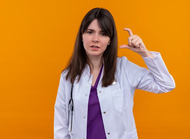 聴診器で医療ローブでイライラする若い女性医師は、コピースペースと孤立したオレンジ色の背景に指で何かを保持するふりをします