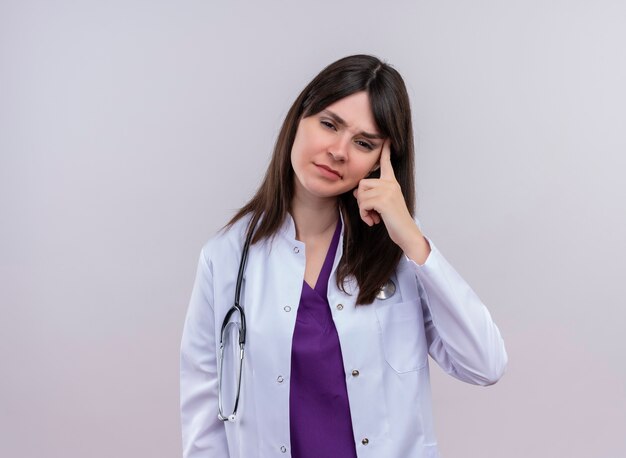 Annoyed young female doctor in medical robe with stethoscope puts finger to her temple on isolated white background with copy space