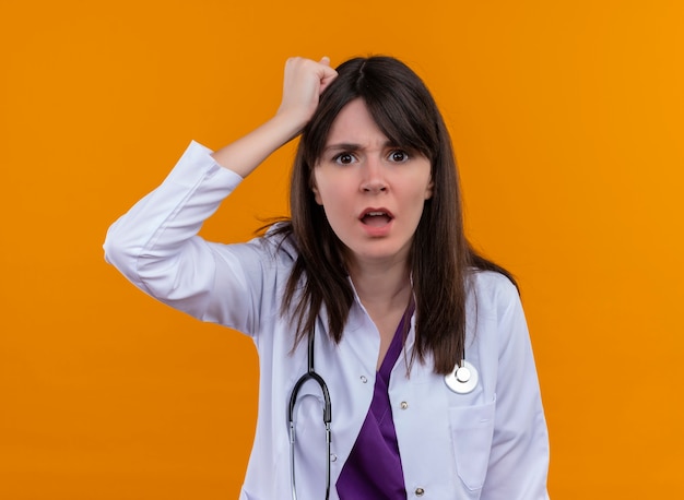 Annoyed young female doctor in medical robe with stethoscope puts fist on head on isolated orange background
