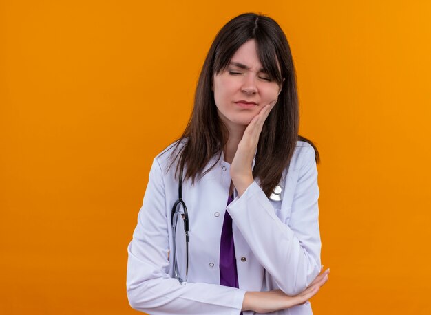 聴診器と医療ローブでイライラする若い女性医師は、コピースペースと孤立したオレンジ色の背景に頬に手を置きます