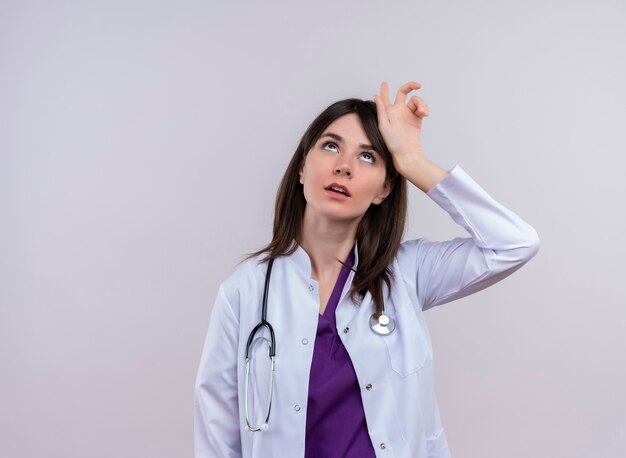 Annoyed young female doctor in medical robe with stethoscope puts hand on head and looks up on isolated white background with copy space