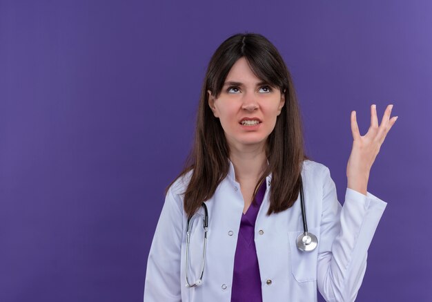 Annoyed young female doctor in medical robe with stethoscope raises hand up on isolated violet background with copy space