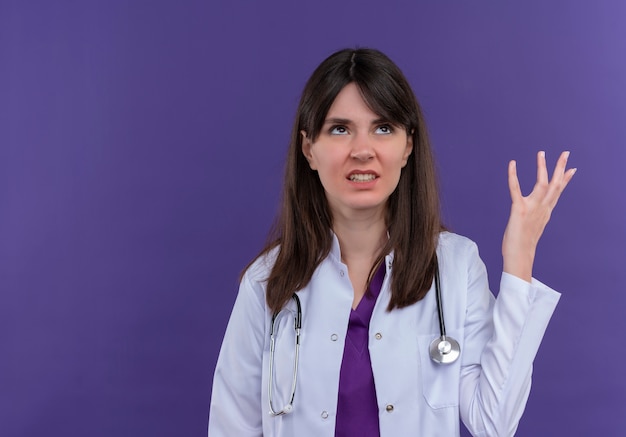 Раздраженная молодая женщина-врач в медицинском халате со стетоскопом поднимает руку на изолированном фиолетовом фоне с копией пространства
