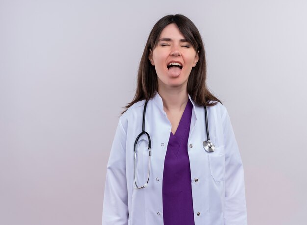 Annoyed young female doctor in medical robe with stethoscope with closed eyes stucks out tongue on isolated white background with copy space