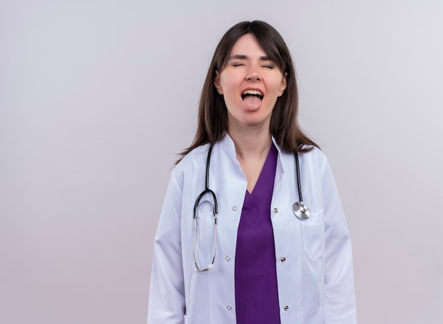 Раздраженная молодая женщина-врач в медицинском халате со стетоскопом с закрытыми глазами высовывает язык на изолированном белом фоне с копией пространства
