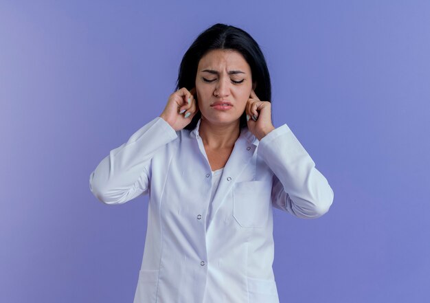 Annoyed young female doctor wearing medical robe putting hands in ears with closed eyes 