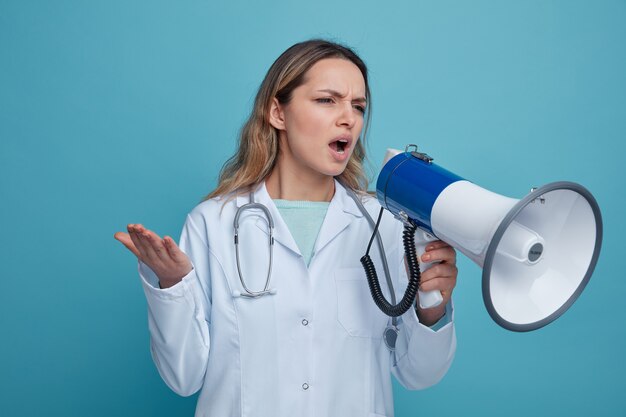 Annoyed young female doctor wearing medical robe and stethoscope around neck looking at side talking by speaker showing empty hand 