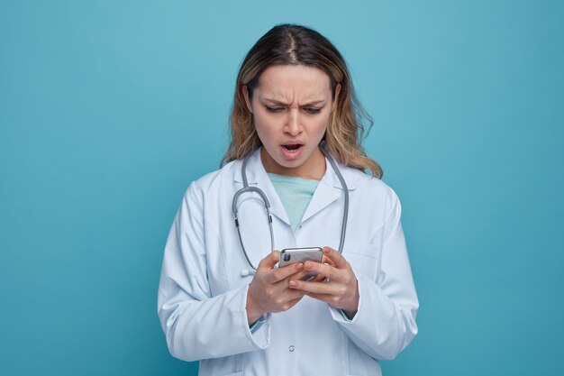 Раздраженная молодая женщина-врач в медицинском халате и стетоскопе на шее, используя свой мобильный телефон