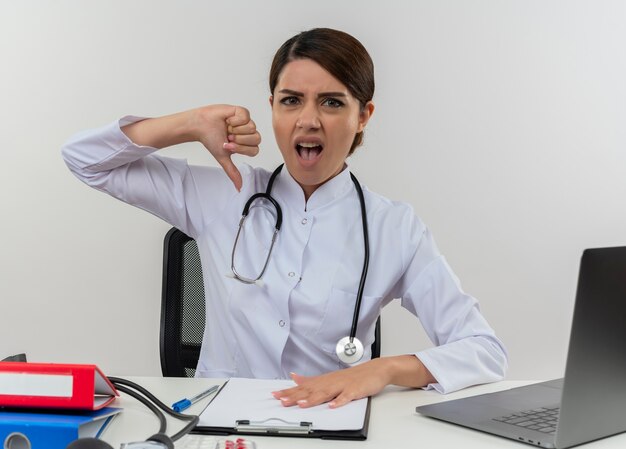 Annoyed young female doctor wearing medical robe and stethoscope sitting at desk with medical tools and laptop showing thumb down isolated on white wall