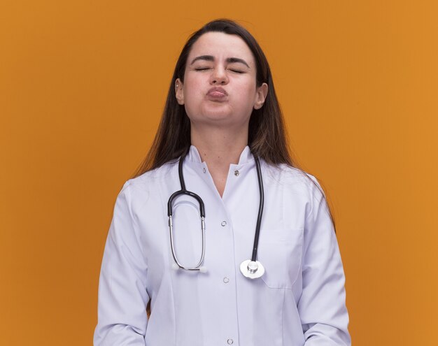 Annoyed young female doctor wearing medical robe with stethoscope blows cheeks and stands with closed eyes isolated on orange wall with copy space