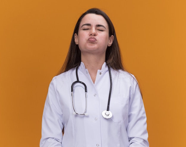 Раздраженная молодая женщина-врач в медицинском халате со стетоскопом ударяет по щекам и стоит с закрытыми глазами, изолированными на оранжевой стене с копией пространства