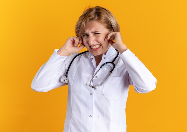 オレンジ色の壁に隔離された聴診器の閉じた耳と医療ローブを身に着けているイライラする若い女性医師