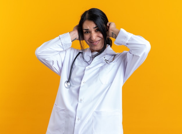 Annoyed young female doctor wearing medical robe with stethoscope covered ears isolated on yellow wall