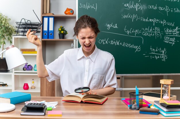 annoyed young female math teacher taking off glasses sitting at desk with school supplies holding magnifying glass keeping hand on open book screaming with closed eyes in classroom